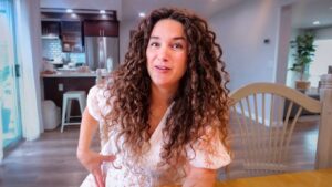 Woman with long curly hair sits at a wooden dining table in a bright kitchen, smiling at the camera. (Informative, concise)