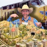 Man in a cowboy hat and blue plaid shirt points to a detailed model railroad landscape with tunnels and a church, set in front of a large yellow locomotive behind him.