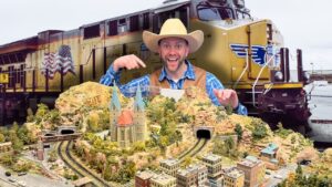 Man in a cowboy hat and blue plaid shirt points to a detailed model railroad landscape with tunnels and a church, set in front of a large yellow locomotive behind him.