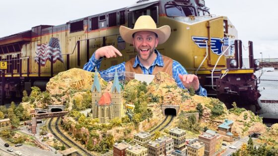 Man in a cowboy hat and blue plaid shirt points to a detailed model railroad landscape with tunnels and a church, set in front of a large yellow locomotive behind him.
