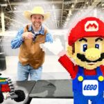 Smiling man in a cowboy hat and brown vest points at a giant LEGO Mario statue beside a colorful LEGO car at a convention hall.