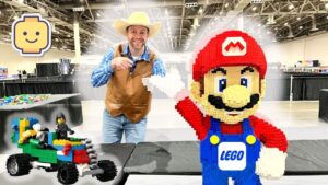 Smiling man in a cowboy hat and brown vest points at a giant LEGO Mario statue beside a colorful LEGO car at a convention hall.