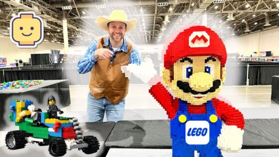 Smiling man in a cowboy hat and brown vest points at a giant LEGO Mario statue beside a colorful LEGO car at a convention hall.