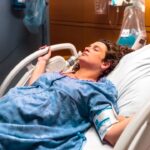 Woman in a blue hospital gown resting in a hospital bed with a blood pressure cuff on her arm, in a patient room.