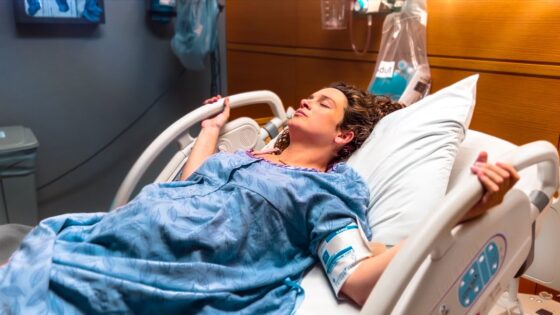 Woman in a blue hospital gown resting in a hospital bed with a blood pressure cuff on her arm, in a patient room.