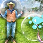 Man in a cowboy hat and vest poses on a grassy field, surrounded by giant bubbles and a cartoon boy drawing bubbles on the right side.