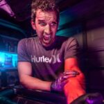 Man wearing a Hurley t-shirt grimaces as he leans over an open car engine in a dim, neon-lit garage.