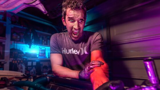 Man wearing a Hurley t-shirt grimaces as he leans over an open car engine in a dim, neon-lit garage.
