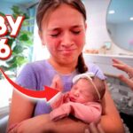 Mother cradles a newborn baby wearing pink in a cozy room, with bold 'BABY #6' text overlay and a hand reaching toward the baby.