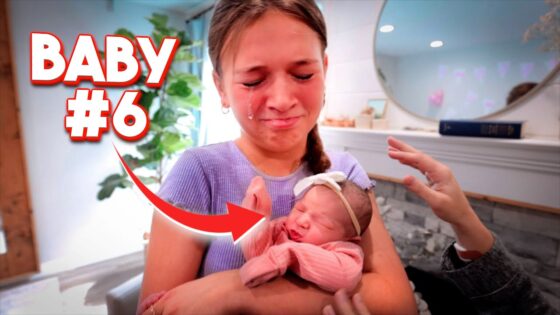 Mother cradles a newborn baby wearing pink in a cozy room, with bold 'BABY #6' text overlay and a hand reaching toward the baby.