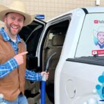 Smiling man in a cowboy hat and brown vest points at a white van door, holding a blue vacuum hose beside a festive setup.