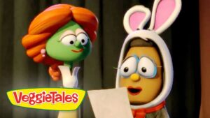 Two VeggieTales characters on a stage: a green vegetable with orange hair speaks to a brown character wearing bunny ears, holding a sheet of paper. Logo reads VeggieTales in the corner.