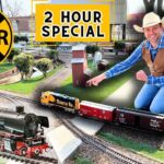 Man kneeling beside a detailed model railroad layout with trains, tracks, and miniature buildings, under a bright '2 HOUR SPECIAL' sign and a large yellow RR crossing symbol.