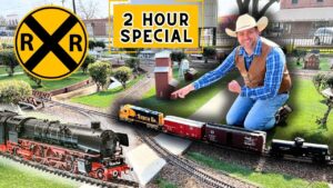 Man kneeling beside a detailed model railroad layout with trains, tracks, and miniature buildings, under a bright '2 HOUR SPECIAL' sign and a large yellow RR crossing symbol.