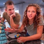 Two people lean over a countertop, talking, beside a tall macaron tower in pastel colors inside a kitchen.