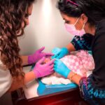 Two healthcare workers wearing gloves and masks tend to a newborn baby in a hospital bassinet, wrapped in a pink blanket, during a medical checkup