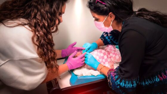 Two healthcare workers wearing gloves and masks tend to a newborn baby in a hospital bassinet, wrapped in a pink blanket, during a medical checkup