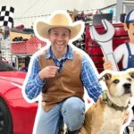 Man in a cowboy hat and vest kneels beside a large dog in a garage setting with a red sports car and racing checkered flags in the background.