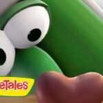 Close-up of a green VeggieTales character with large googly eyes, looking down at a brown object on a wooden surface, VeggieTales logo in the corner.