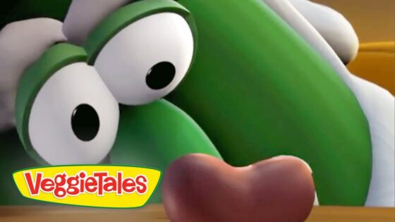 Close-up of a green VeggieTales character with large googly eyes, looking down at a brown object on a wooden surface, VeggieTales logo in the corner.