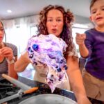 Three people gather around a kitchen counter, holding a colorful purple-blue ice sculpture head they made together.