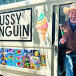Smiling man in a cowboy hat and brown vest stands at the open door of an ice cream truck reading 'The Fussy Penguin Company', holding two ice cream treats.