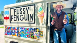 Smiling man in a cowboy hat and brown vest stands at the open door of an ice cream truck reading 'The Fussy Penguin Company', holding two ice cream treats.