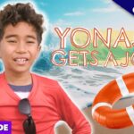 Smiling boy in a red shirt at the beach with an orange lifebuoy and the headline 'YONAS GETS A JOB'.