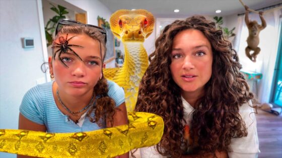 Two young women indoors posing with a large yellow snake draped across the foreground, looking at the camera.