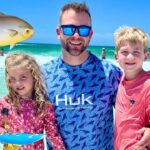 Family at the beach: father with two kids posing in the surf with cartoon sea creatures and a fishing net overlay nearby. coast and turquoise water in the background.