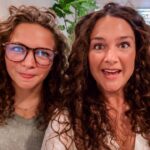 Two women with curly hair posing for a selfie in a cozy living room setting with lounge chairs in the background, smiling at the camera.