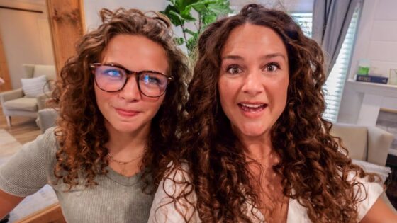 Two women with curly hair posing for a selfie in a cozy living room setting with lounge chairs in the background, smiling at the camera.