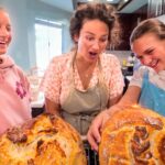 Three friends in a bright kitchen smile and laugh while admiring two large loaves of bread cooling on a rack on the counter behind them.