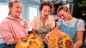 Three friends in a bright kitchen smile and laugh while admiring two large loaves of bread cooling on a rack on the counter behind them.