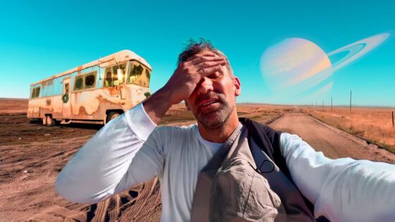 Man taking a selfie on a dusty desert road; an old rusted bus sits to his left with a giant ringed planet in the bright blue sky behind him.