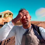 Selfie of a man on a dusty rural road, with an abandoned rusty bus to the left and a giant ringed planet in the blue sky behind him.