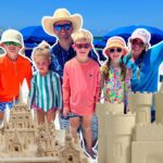 Group of people on a sunny beach posing with intricate sandcastles and blue umbrellas in the background.