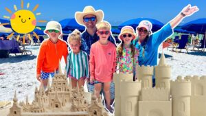 Group of people on a sunny beach posing with intricate sandcastles and blue umbrellas in the background.