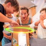A woman and three kids cheer as they open a cardboard box with a glowing light inside on a living room table.