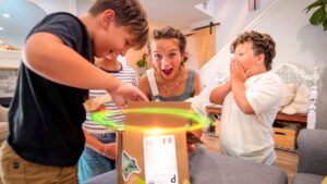 A woman and three kids cheer as they open a cardboard box with a glowing light inside on a living room table.