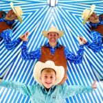 Group of five people dressed as cowboys with brown vests, blue shirts, and hats, standing under a blue-and-white striped canopy with arms outstretched.