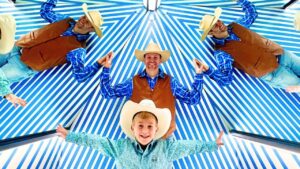 Group of five people dressed as cowboys with brown vests, blue shirts, and hats, standing under a blue-and-white striped canopy with arms outstretched.