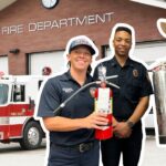 Firefighters and a safety instructor stand with fire extinguishers in front of a fire station.