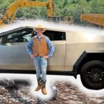 Man in a cowboy hat and vest stands beside a silver futuristic pickup on a rocky construction site.