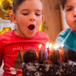 Three children lean in as they blow out lit candles on a chocolate birthday cake
