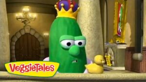 Animated green cucumber with a yellow crown, looking concerned in a castle interior beside two ducklings, from VeggieTales logo visible nearby