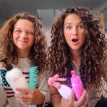 Two girls with curly hair sit on a living room floor, holding assorted hair-care products and brushes for a hair-tutorial video.