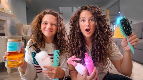 Two girls with curly hair sit on a living room floor, holding assorted hair-care products and brushes for a hair-tutorial video.