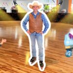 Man in a cowboy vest and hat stands on inline skates in an indoor roller-skating rink; a cartoon kid on skates is on the left and blue roller skates appear on the right.