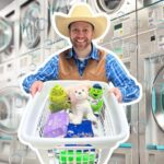 Smiling man in a cowboy hat and vest pushes a laundry basket filled with stuffed animals and laundry supplies in a laundromat.